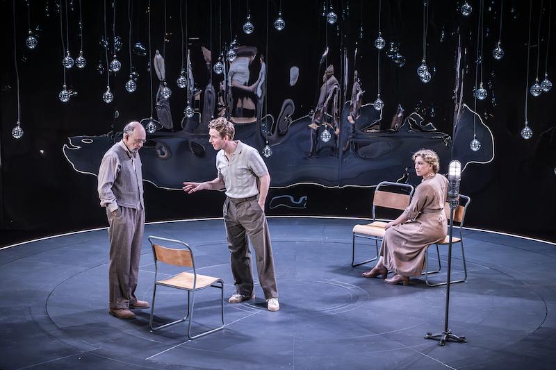 Richard Schiff and Damien Molony standing and speaking to each other, as Alex Kingston sits and listens