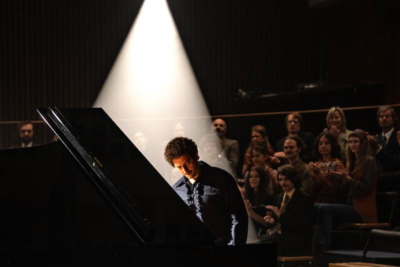 John Magaro as Keith Jarrett, sitting at a piano in a darkened room, with audience behind 